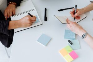 The hands of two people write on sticky notes and in notebooks.