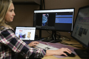 A woman works in front of a monitor and laptop, using Genna’s VidGen tool to produce social media videos.